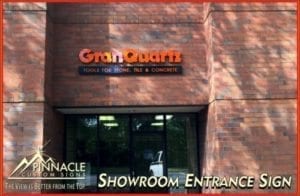 Exterior view of a brick building with the business name 'GranQuartz' displayed in orange channel letters above the entrance. Below the name, the tagline 'Tools for Stone, Tile & Concrete' is visible. The sign is mounted above large glass doors. The Pinnacle Custom Signs logo and tagline 'The View is Better from the Top' are in the bottom left corner of the image. The text 'Showroom Entrance Sign' is in the bottom right corner.