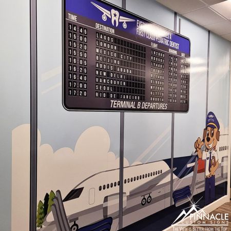 Window graphics for First Class Pediatric Dentist, featuring an airport departure board with various destinations and an illustration of an airplane and a friendly pilot character, displayed on an interior wall